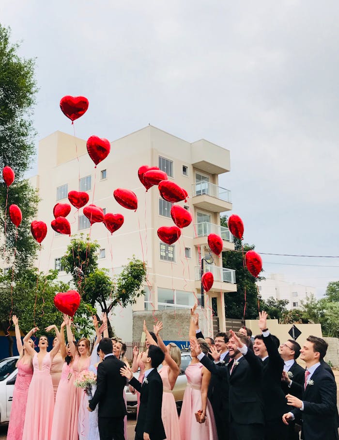 A joyful wedding party celebrating outdoors with red heart balloons in the air against an urban backdrop.